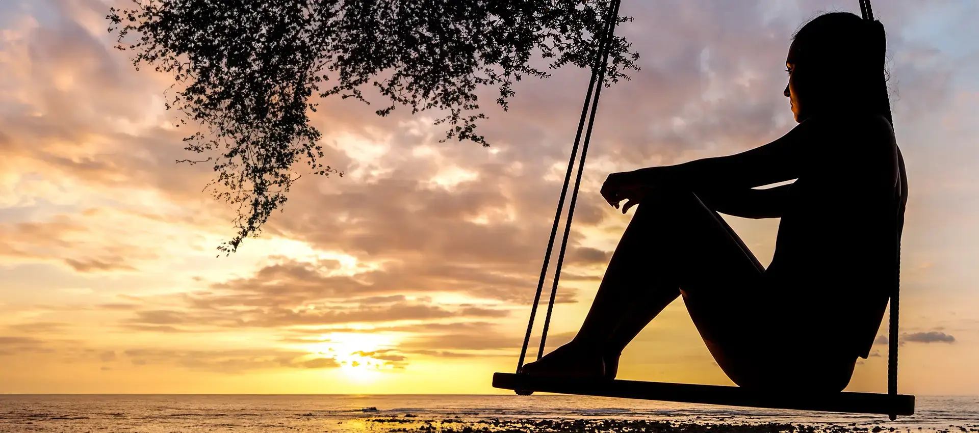 A female sitting on a swing on a swing set looking out at the water and a sunset.