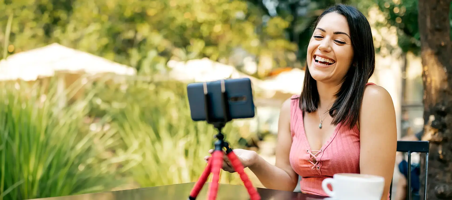 A hispanic woman sitting outside and smiling while having a video call through her smartphone on a tripod.